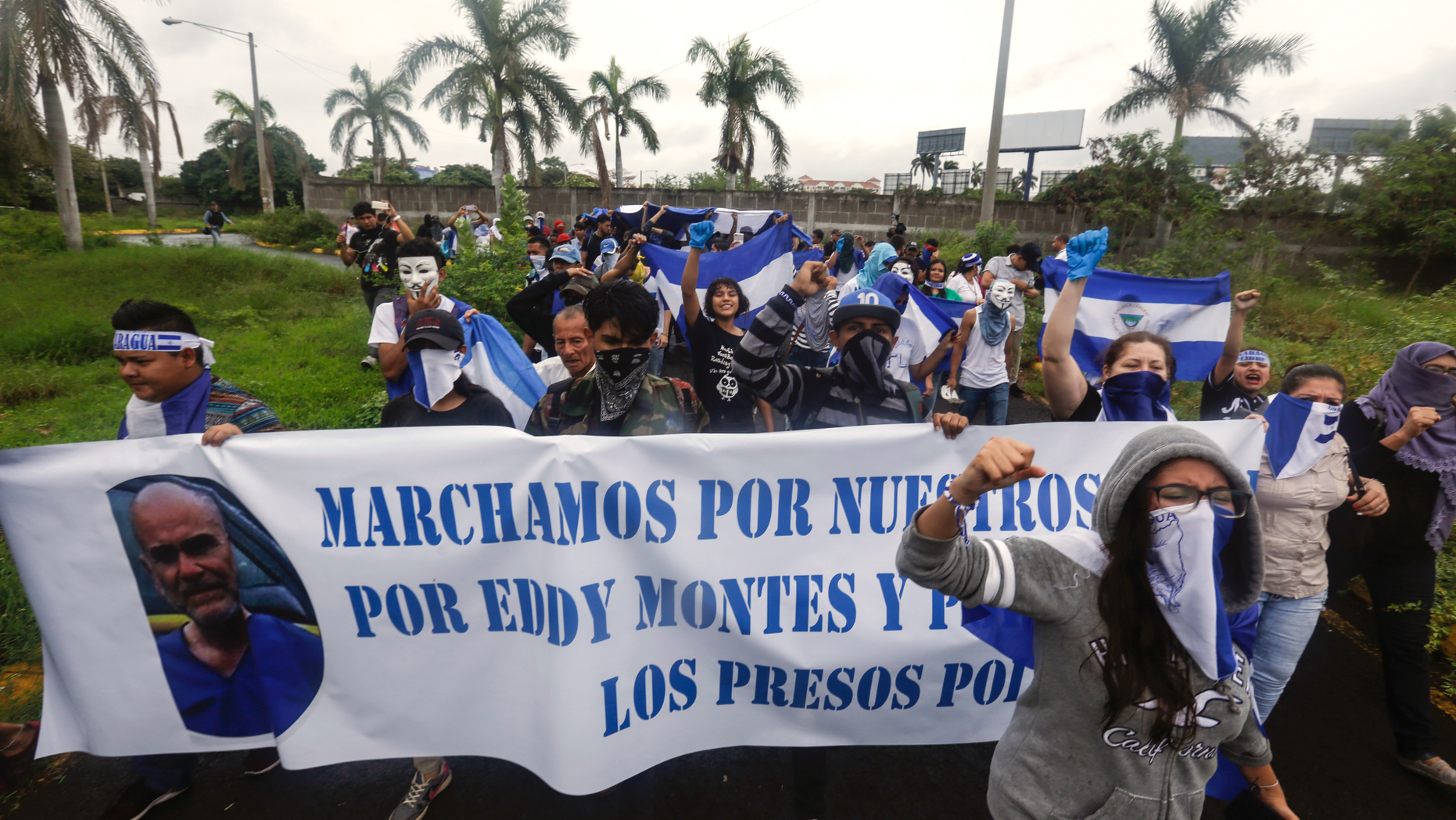 Anti-government protesters, some wearing masks to hide their identity from authorities, protest on the grounds of the Cathedral after their march was blocked by police in Managua, Nicaragua, Sunday, May 26, 2019. The banner features Eddy Montes Praslin, a Nicaraguan-American dual national who was a staunch opponent of the government of President Daniel Ortega and who died in a prison disturbance in Nicaragua. (AP Photo/Alfredo Zuniga)