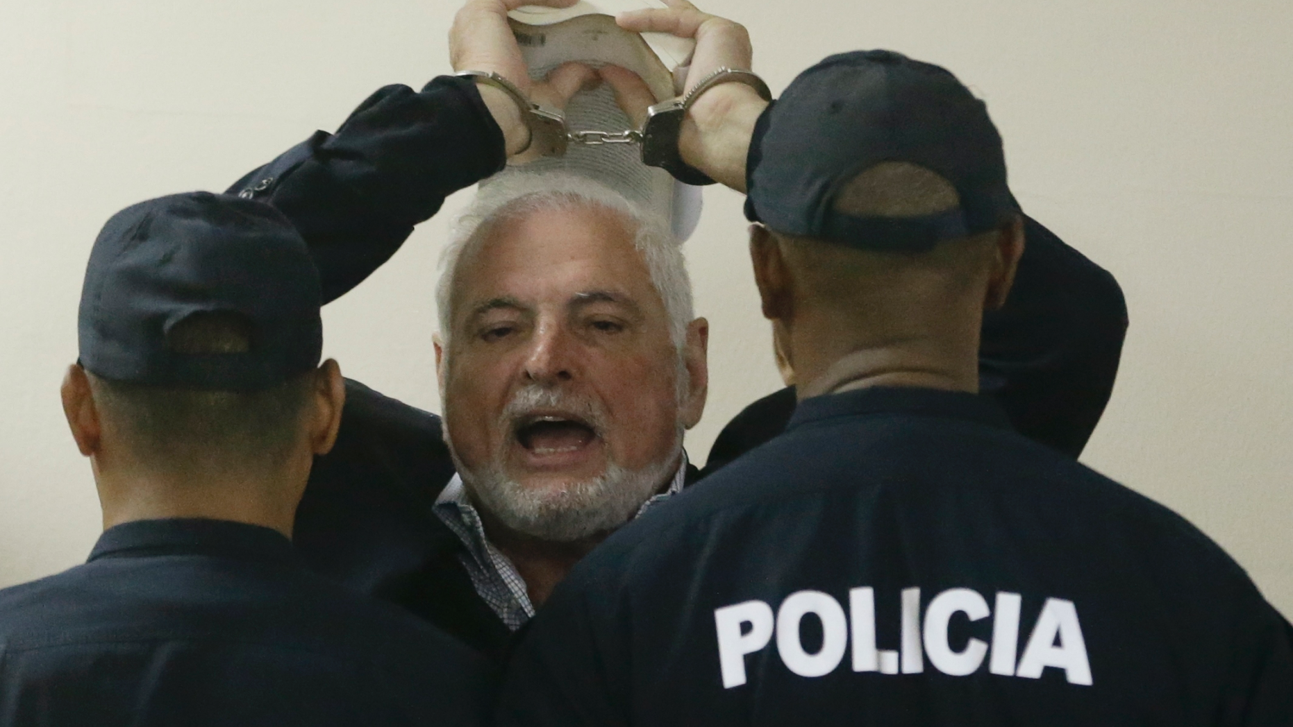 FILE - In this Nov. 19, 2018 file photo, Panama's former President President Ricardo Martinelli answers journalists questions as he raises his handcuffed hands, holding a book, while escorted by police to a hearing at the Supreme Court in Panama City. A Panamanian court has ordered on Wednesday, June 12, 2019, that former President Martinelli to be transferred from jail to house arrest. (AP Photo/Arnulfo Franco, File)