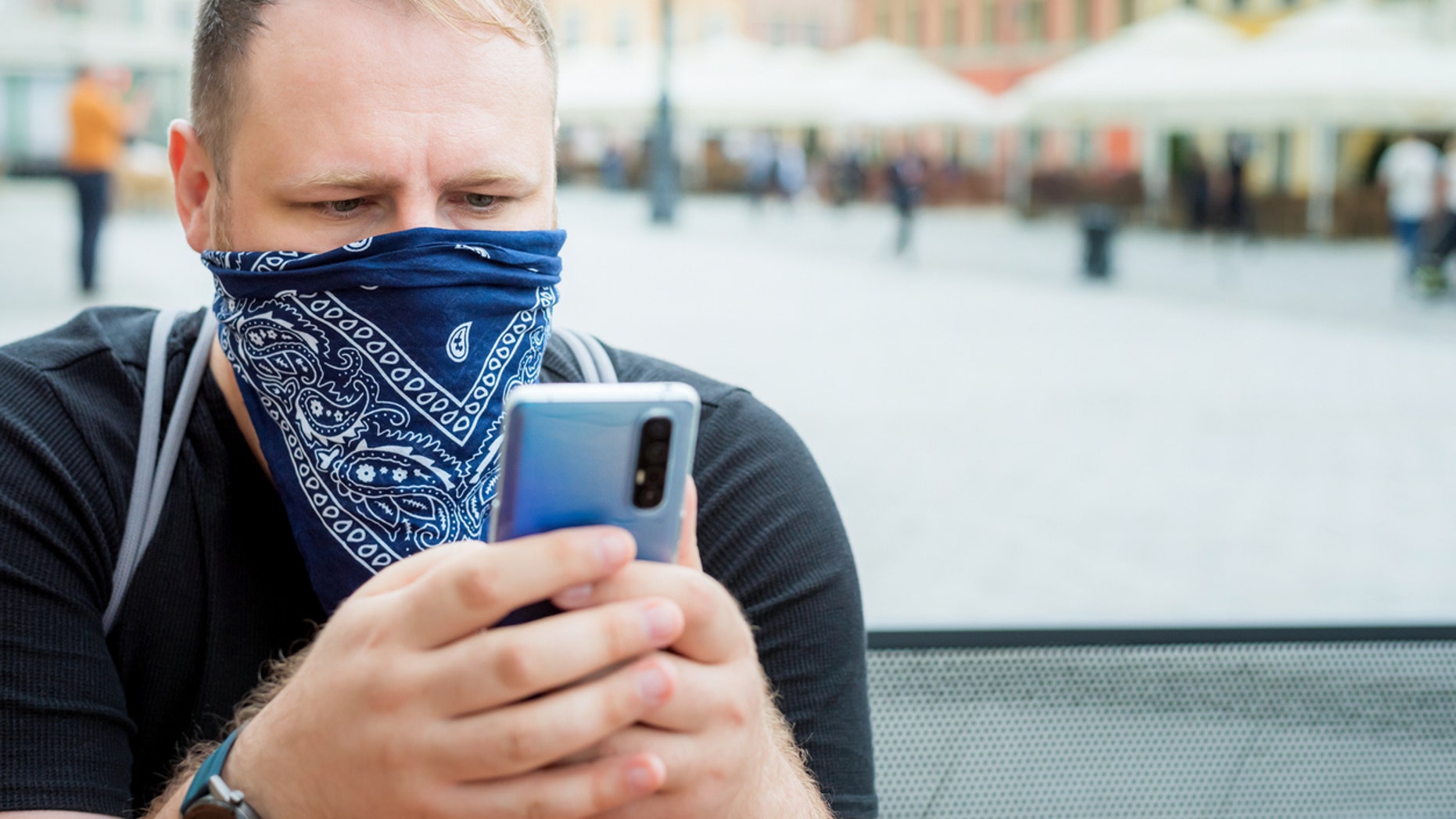 Adult man in a bandana as a face mask uses a smartphone in the city cafe terrace. 