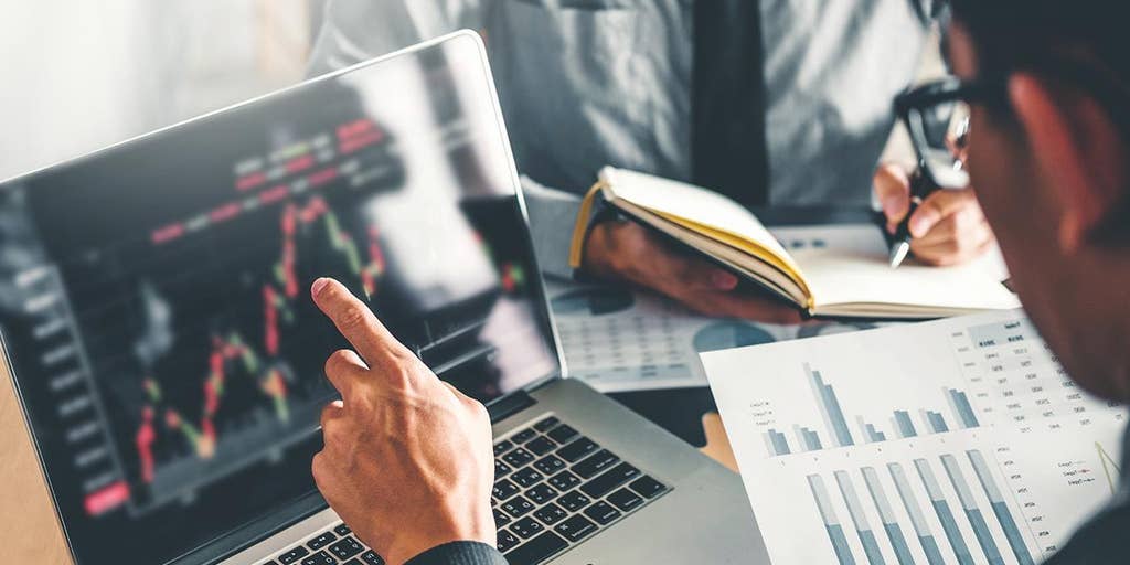  A person is pointing at a laptop screen displaying stock market charts while another person is taking notes, with a notebook and pen in hand.