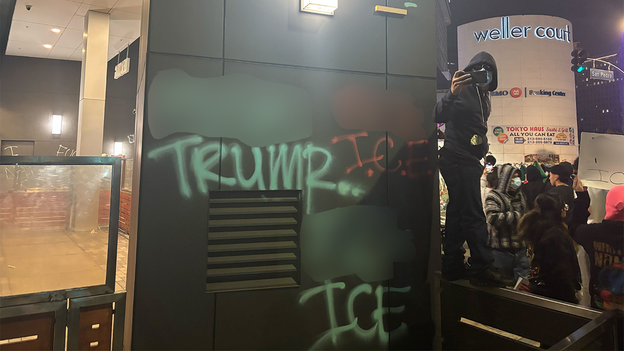 A masked protestor films from atop a ledge next to anti-Trump graffiti