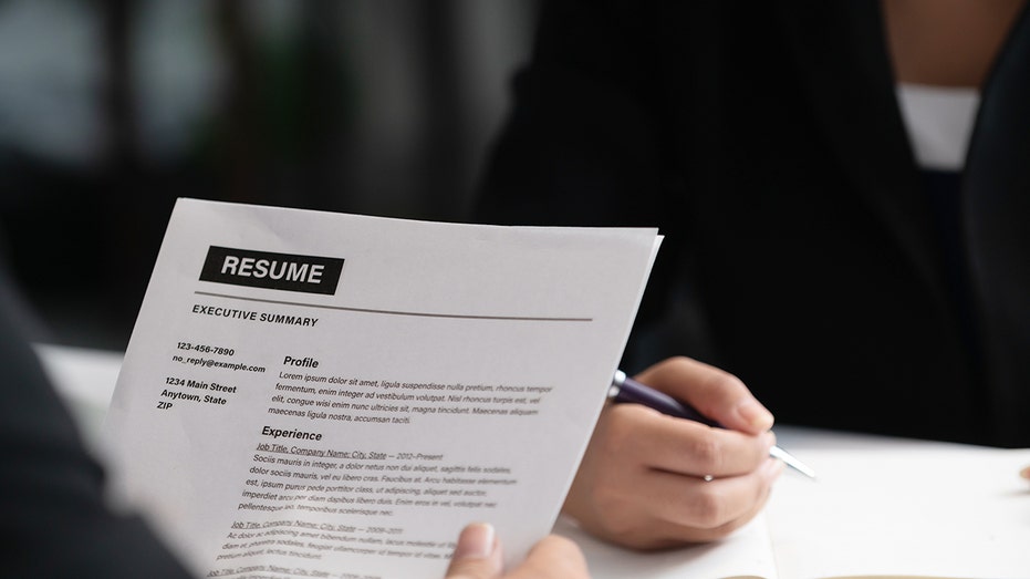 Person holds up resume in front of someone with a pen in-hand