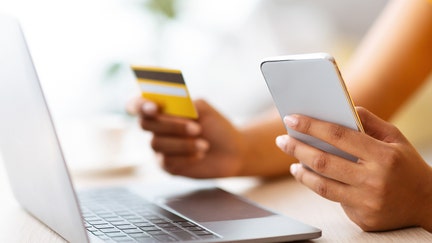 Mobile Banking App. Unrecognizable african american woman holding debit credit card and cellphone, sitting at table with laptop, cheking her online wallet. Blurred background, selective focus - Fox Business News
