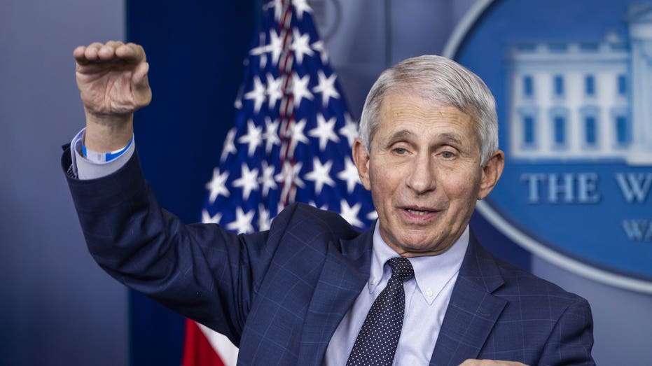 Anthony Fauci in a suit at press conference with right hand in air