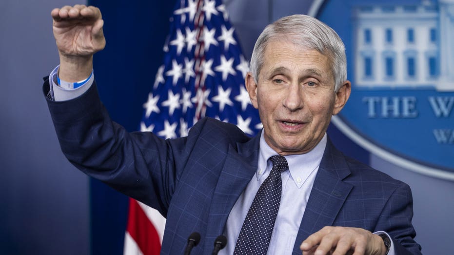 Anthony Fauci in a suit at press conference with right hand in air