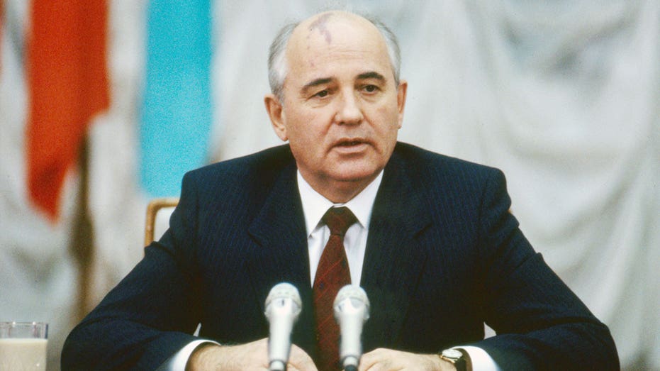 Mikhaill Gorbachev wears a dark suit and sits at a desk speaking into a mic