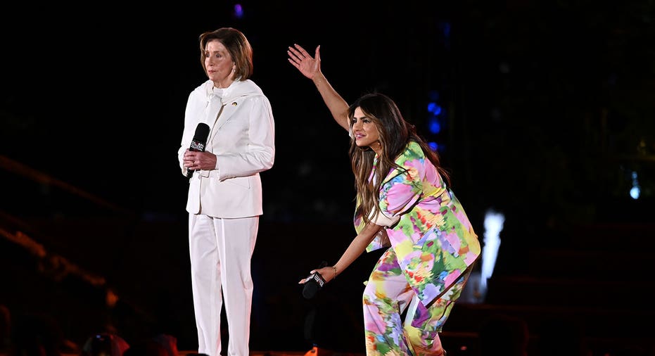 Nancy Pelosi (left) wears white pantsuit while Priyanka Chopra (right) holds up her arms showing off Pelosi