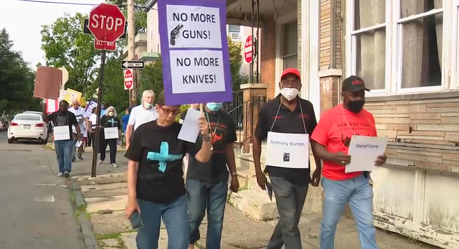 Woman walking with other protesters holding a sign that says 'NO MORE GUNS, NO MORE KNIVES'