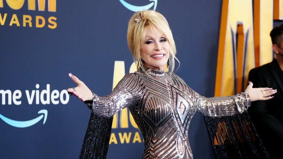 Dolly Parton smiles on the red carpet in a sparkling silver and black dress with tassels on the sleeves