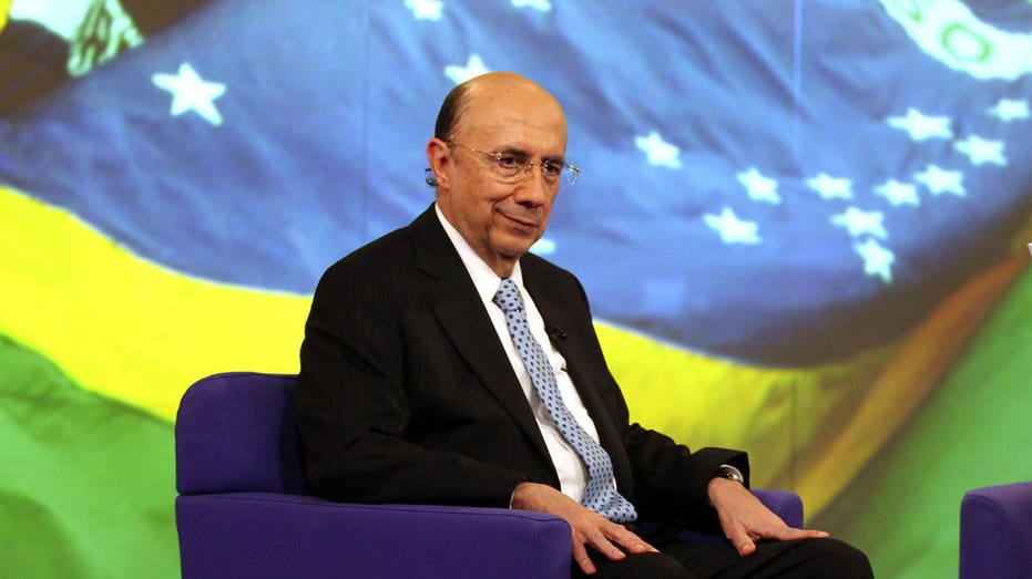 Henrique Mereilles sits in a chair in front of Brazil's flag