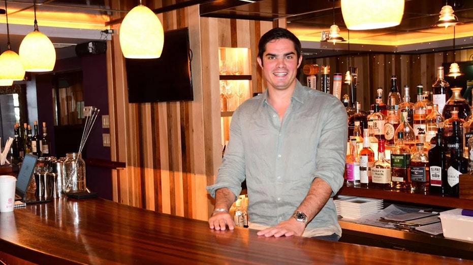 A man in a green button-up shirt stands behind the bar
