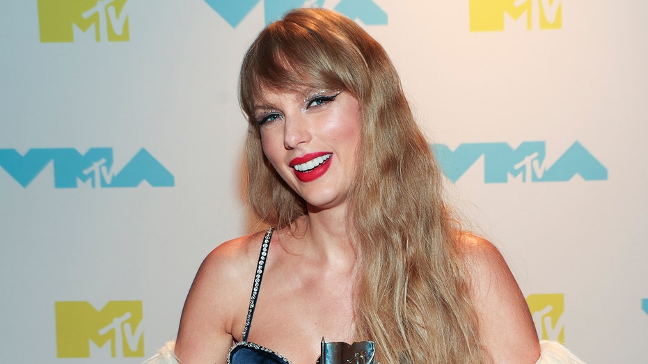 Taylor Swift at the 2022 MTV VIdeo Music Awards smiles in a thin-strapped metallic blue dress with her Moonman awards