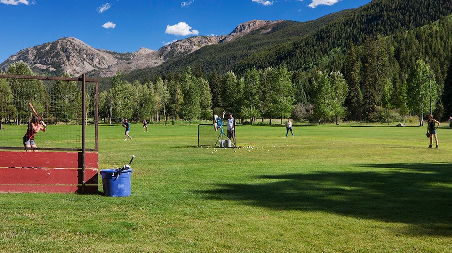 People play baseball as someone pitches behind a net and a batter stands in front of a cage