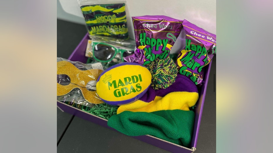 Gift box featuring mardi gras gifts such as sunglasses, purple, yellow and green football, snacks, and a beanie