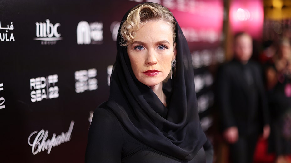 January Jones with a bold red lip stares directly at the camera in a black hooded dress