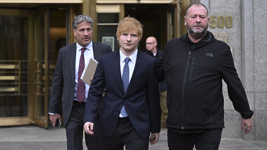 Ed Sheeran walks out of court in New York with a bodyguard and in front of his lawyers wearing a dark suit with a blue tie