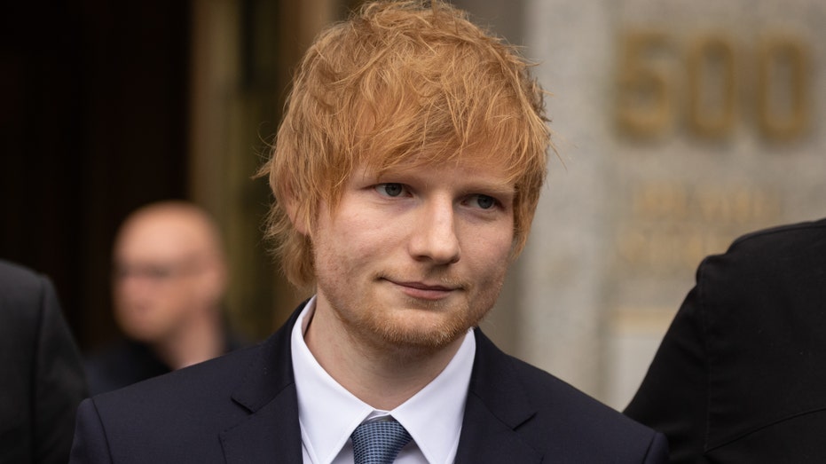 Ed Sheeran looks off in the distance wearing a dark jacket and blue tie with a crisp white button down outside the federal court house in New York