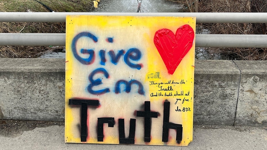 A handmade sign reading 'Give 'em truth' leans against a railing above a water cleanup site in East Palestine, Ohio