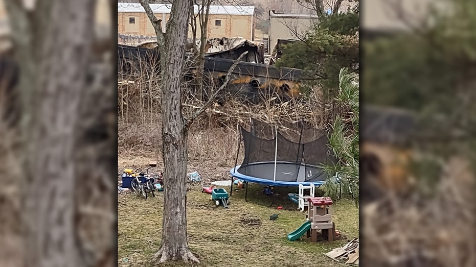 A trampoline and children's toys are scattered across a yard while the wreckage of the East Palestine train derailment can be seen in the background