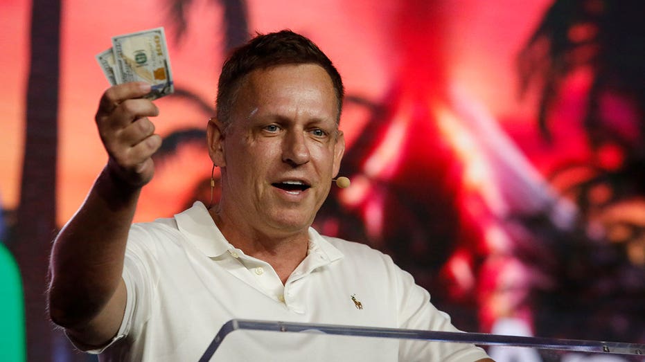 Peter Thiel holds up bills while speaking at a podium