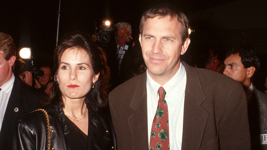 Cindy Silva in black looks off in the distance with husband Kevin Costner in a brown suit jacket and red tie with green flowers, also looking off in the distance (his right) during the "JFK" premiere