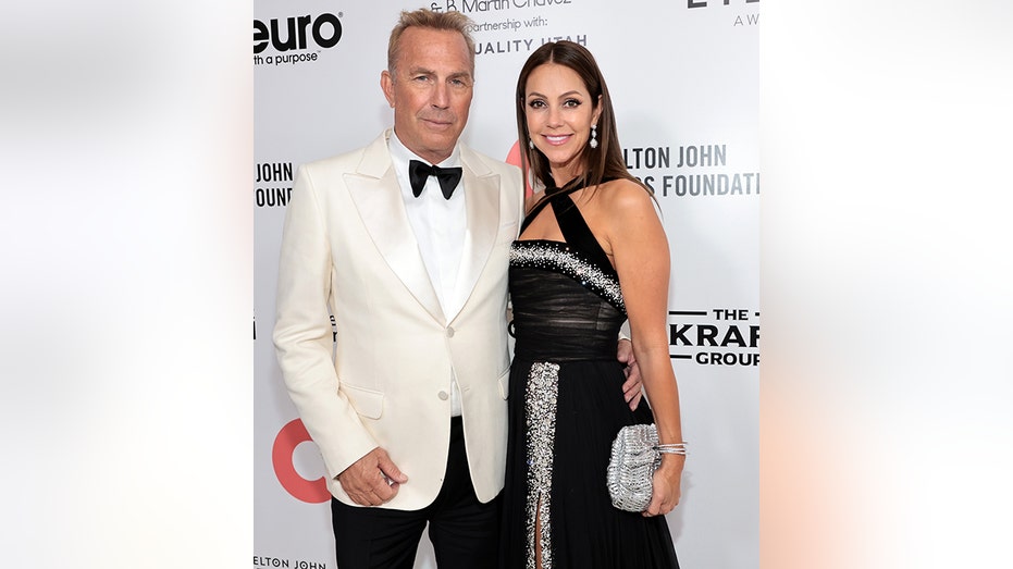 Kevin Costner in a white tuxedo jacket and black pants smiles next to wife Christine in a black criss-cross halter dress with sparkles at Elton John's AIDS Foundation event