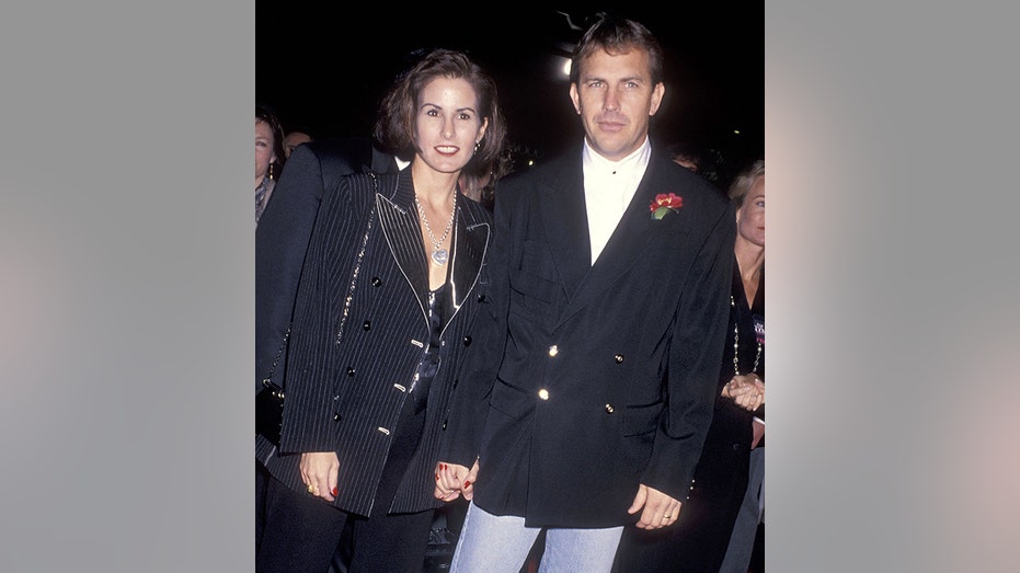 Cindy Silva in a pinstripe blazer holds husband Kevin Costner's hand on the red carpet of the premiere for "The Bodyguard," with Costner in a black suit jacket with a red flower on it