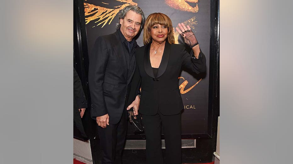 Tina Turner waves wearing a fishnet hand wrap and black suit as her husband Erwin Bach stands next to her at the press night of "Tina: The Tina Turner Musical" in London