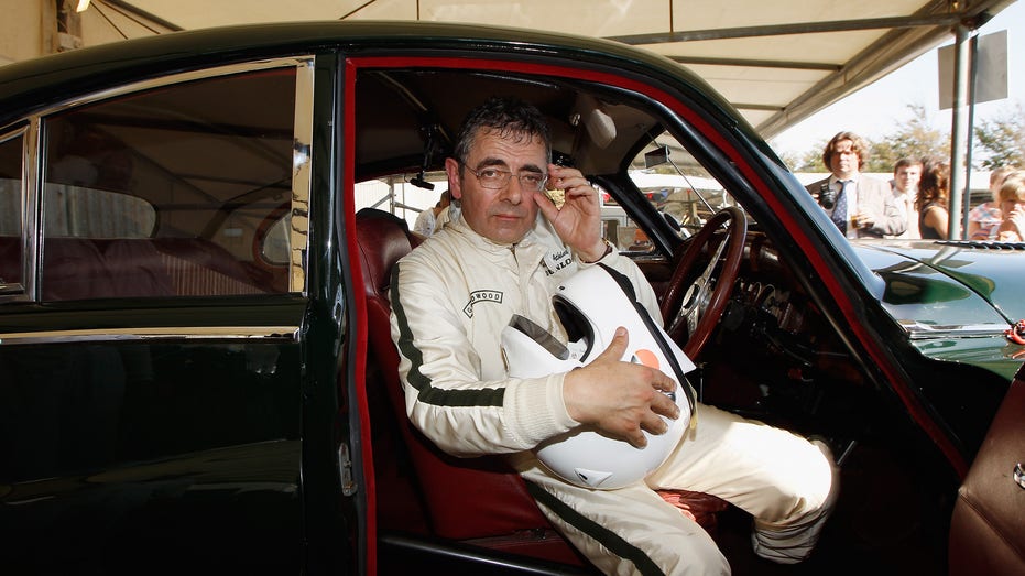 Rowan Atkinson in a racing suit sitting in a car.
