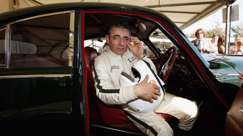 Rowan Atkinson in a racing suit sitting in a car.