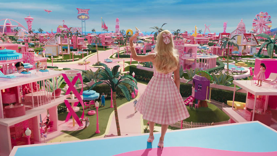Margot Robbie with her back to the camera waves in a plaid pink dress and matching bow over the town of Barbie Land in the movie "Barbie"