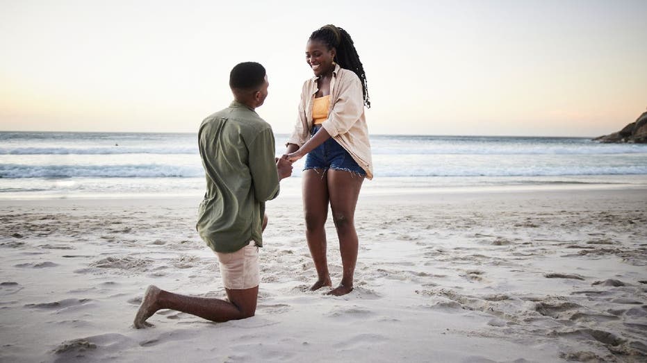 Man proposes marriage to woman on beach. He's down on one knee while he presents an engagement ring.