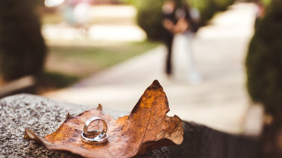 Wedding rings on fallen leaf during autumn while newlyweds pose in background.