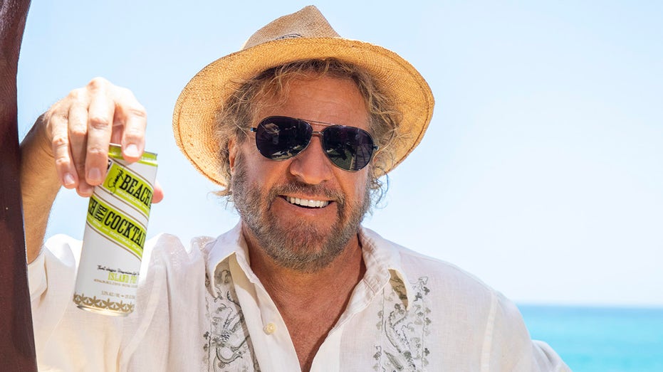 sammy hagar smiling with sunglasses and straw hat holding rum cocktail can in front of ocean