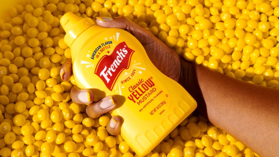 A woman's hand holds a bottle of French's mustard over a backdrop of mustard Skittles
