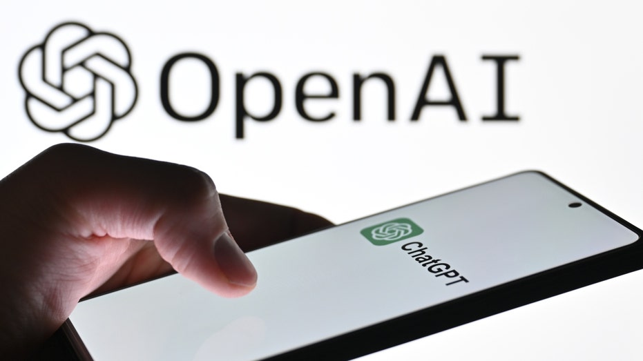 The logo of ChatGPT is displayed on a mobile phone screen in front of a computer screen with the logo of OpenAI