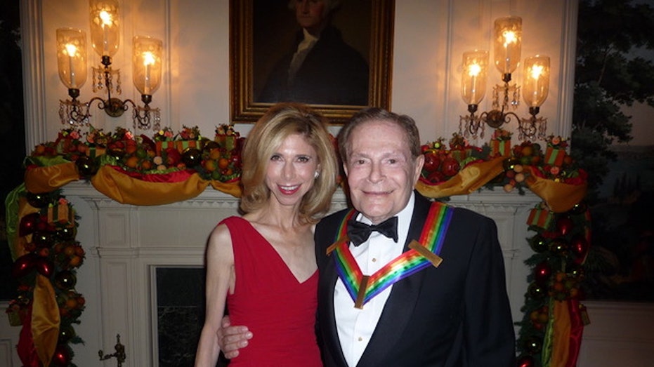 Jane Dorian wearing a red dressing being embraced by Jerr Herman in a suit and bow tie