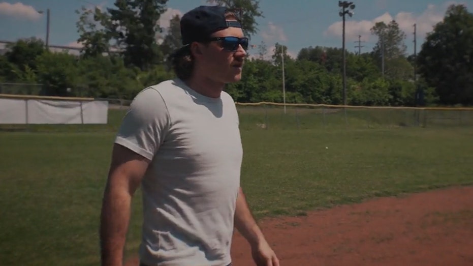 Morgan Wallen wears grey t-shirt and backwards hat on baseball field.