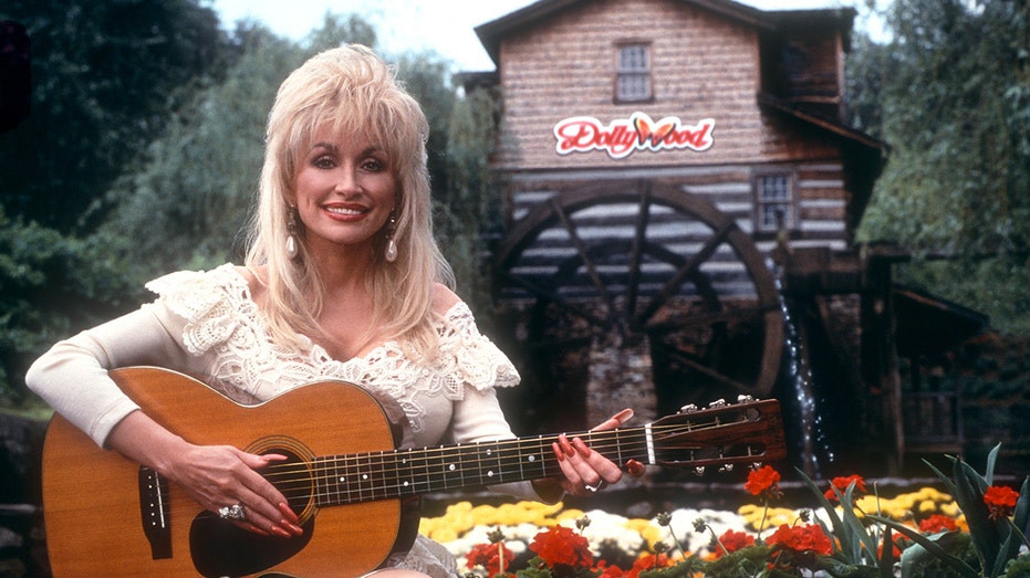 Dolly Parton posing with a guitar in front of a Dollywood sign