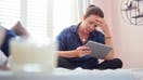 Young woman sits on her bed reading through finances on digital tablet