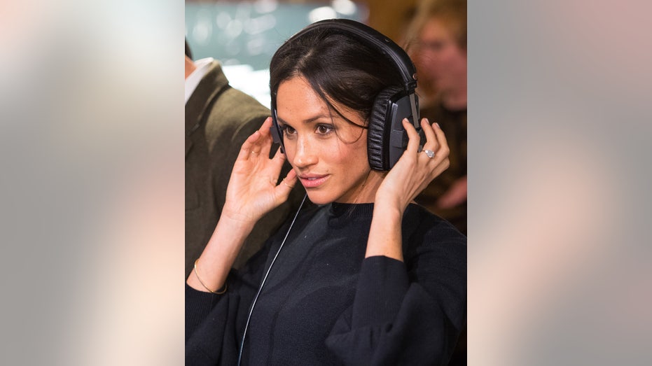 A close-up of Meghan Markle in a black dress listening to her headphones