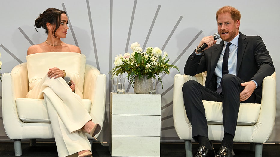 Meghan Markle wearing an ivory sweater and pants sitting next to Prince Harry in a dark suit