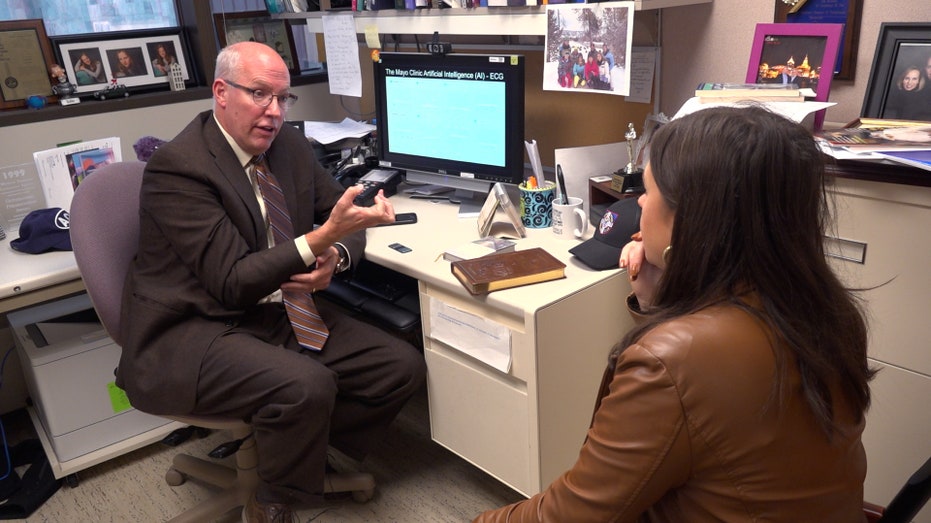 a doctor sits at an office desk explaining an ai ecg screening to a reporter