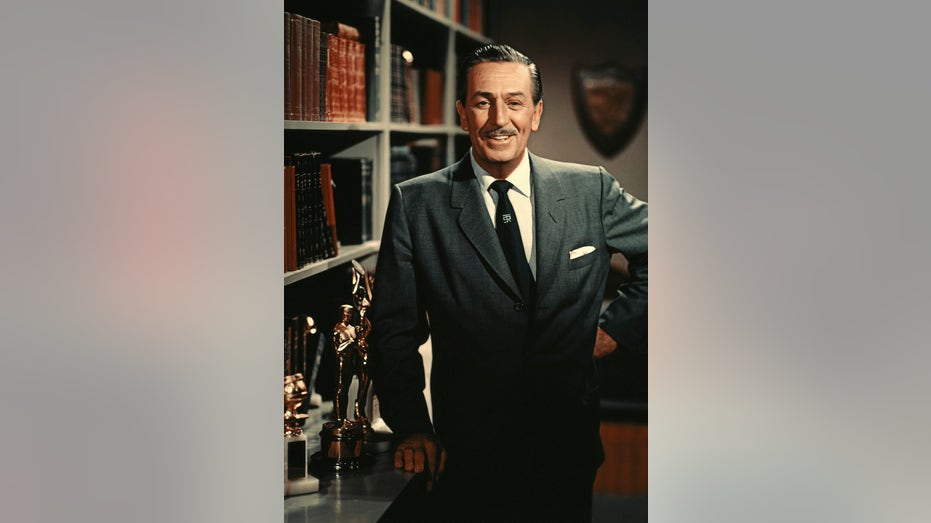 A close-up of Walt Disney smiling next to awards as he wears a suit