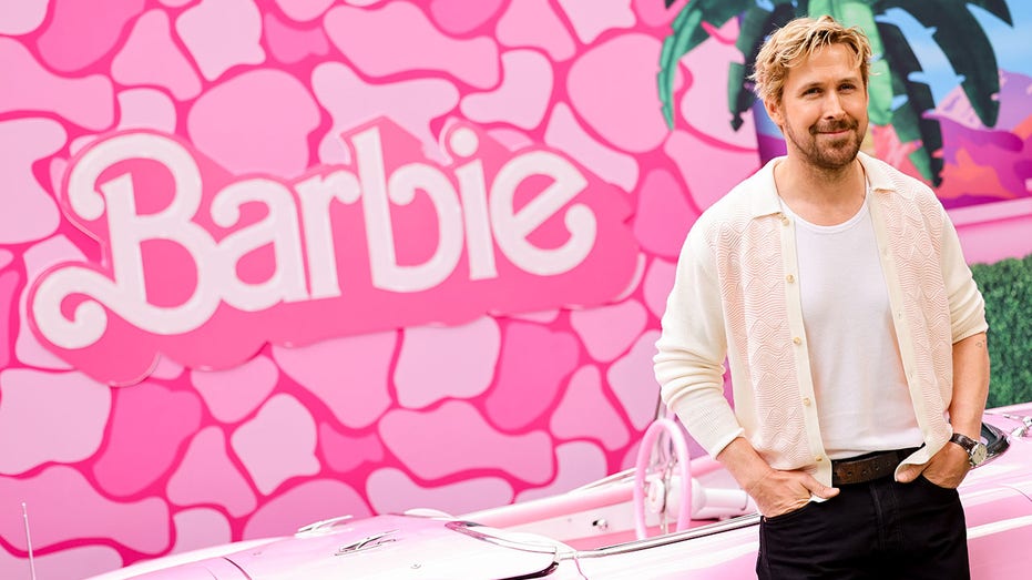 Ryan Gosling in a cream sweater stands in front of a hot pink "Barbie" backdrop