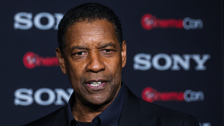 Denzel Washington in a dark suit and dark blue shirt smiles partially on the carpet