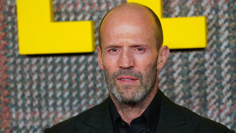 Jason Statham opens his mouth and partially smiles on the carpet in a dark suit and black shirt