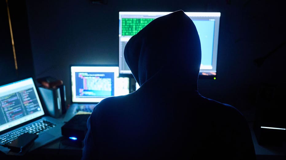 A man types at a computer while fake HTML pages and hacker programs are seen on screens in front of him.