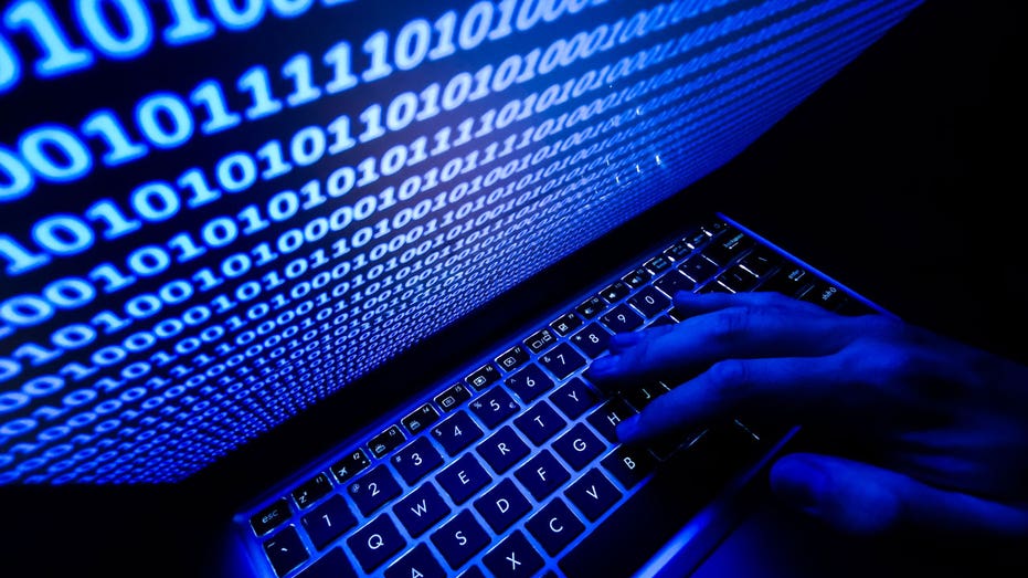 A hand rests on a keyboard, with binary code displayed on a laptop screen.