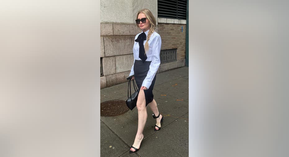 Anna Delvey wears a skirt and shirt with a handbag with an ankle monitor on her left leg